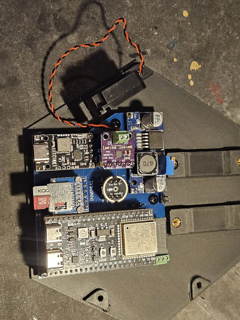 The blue PCB assembly lowered into the black 3D-printed bottom case, brass heat-set inserts and screw posts visible at the corners, with a separate black module and its twisted red-and-black lead pair resting loose above the case, not yet secured