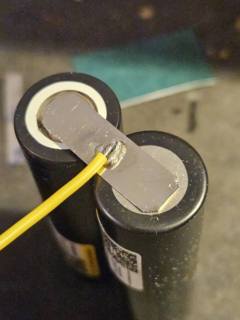 The welded nickel strip bridging the two cell terminals with the balance tap wire soldered at the series junction