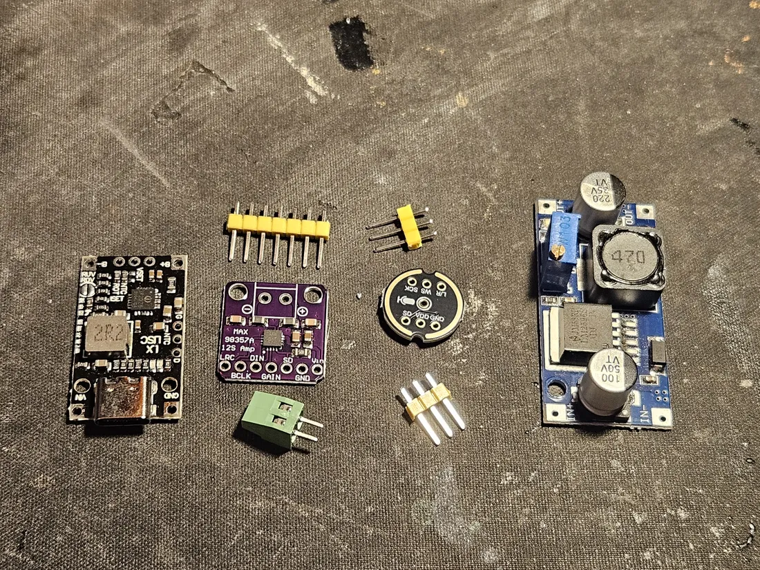 Loose modules laid out: a USB-C charger board, a purple MAX98357A amp, a blue DC-DC LM2596 buck converter, a round INMP441 mic, a green screw terminal, and male header strips