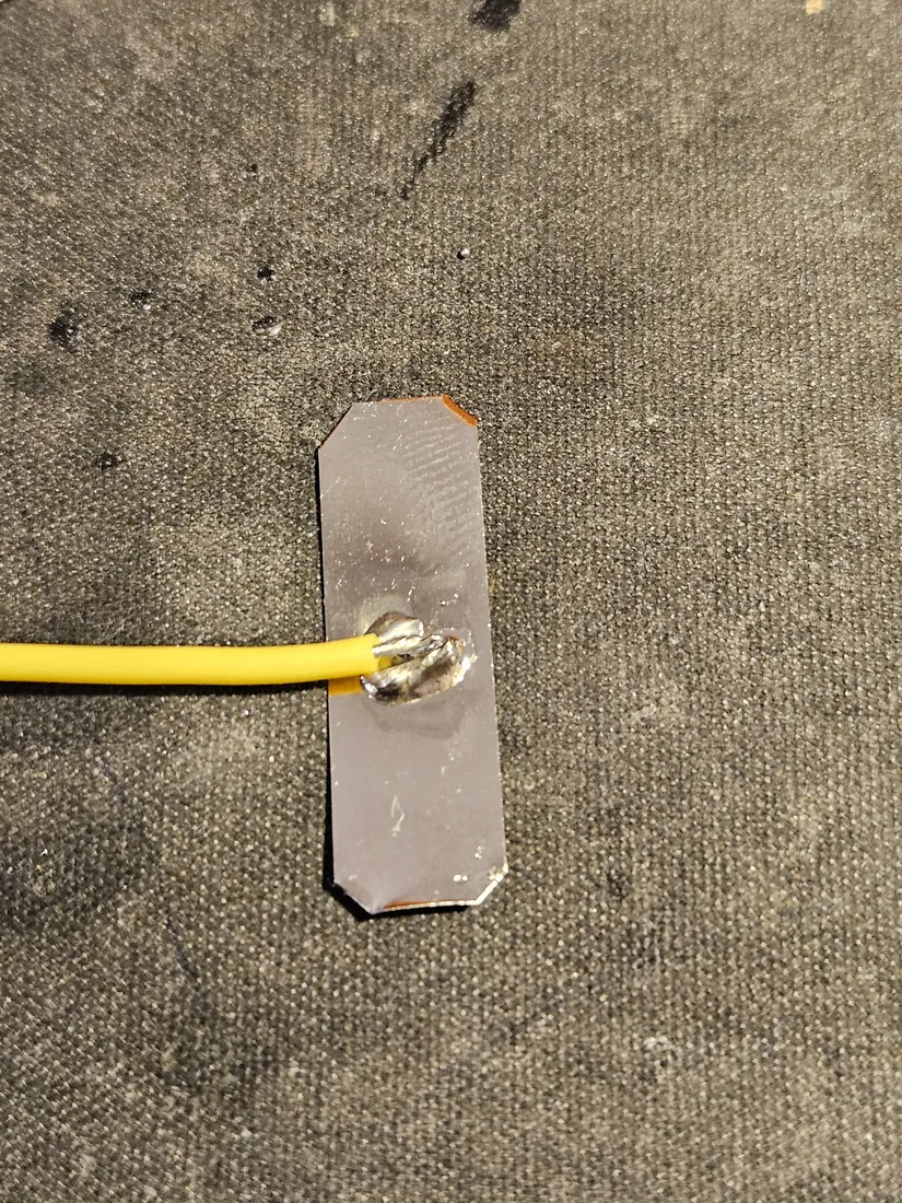 A nickel strip with a yellow lead wire soldered to a tinned pad in its middle