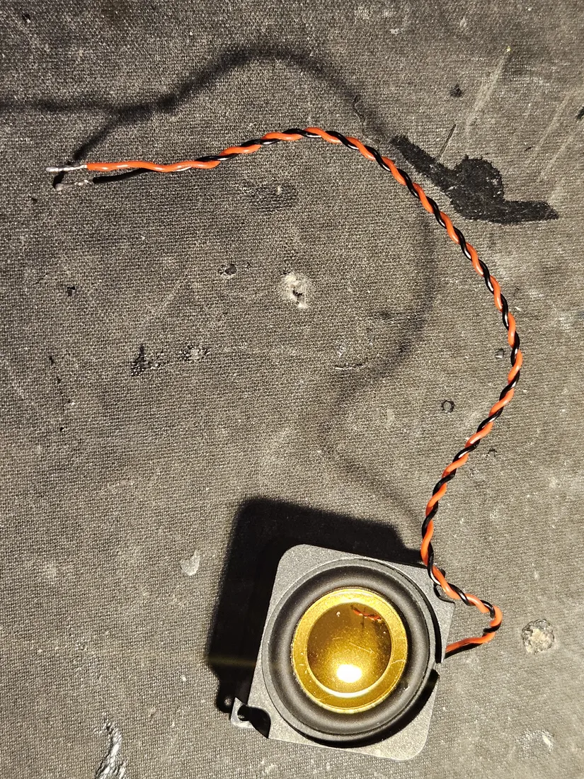 Small round 4 ohm speaker in a square frame with a gold cone and a twisted red-and-black lead pair, ends tinned