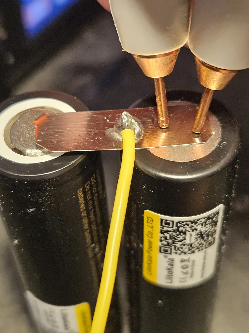 A spot welder pressing two copper electrodes onto a nickel strip laid across the top of a LiitoKala cell, joining it to the neighbouring cell